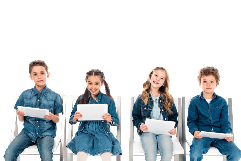 four smiling kids in denim clothes sitting on chairs and using digital tablets isolated on white