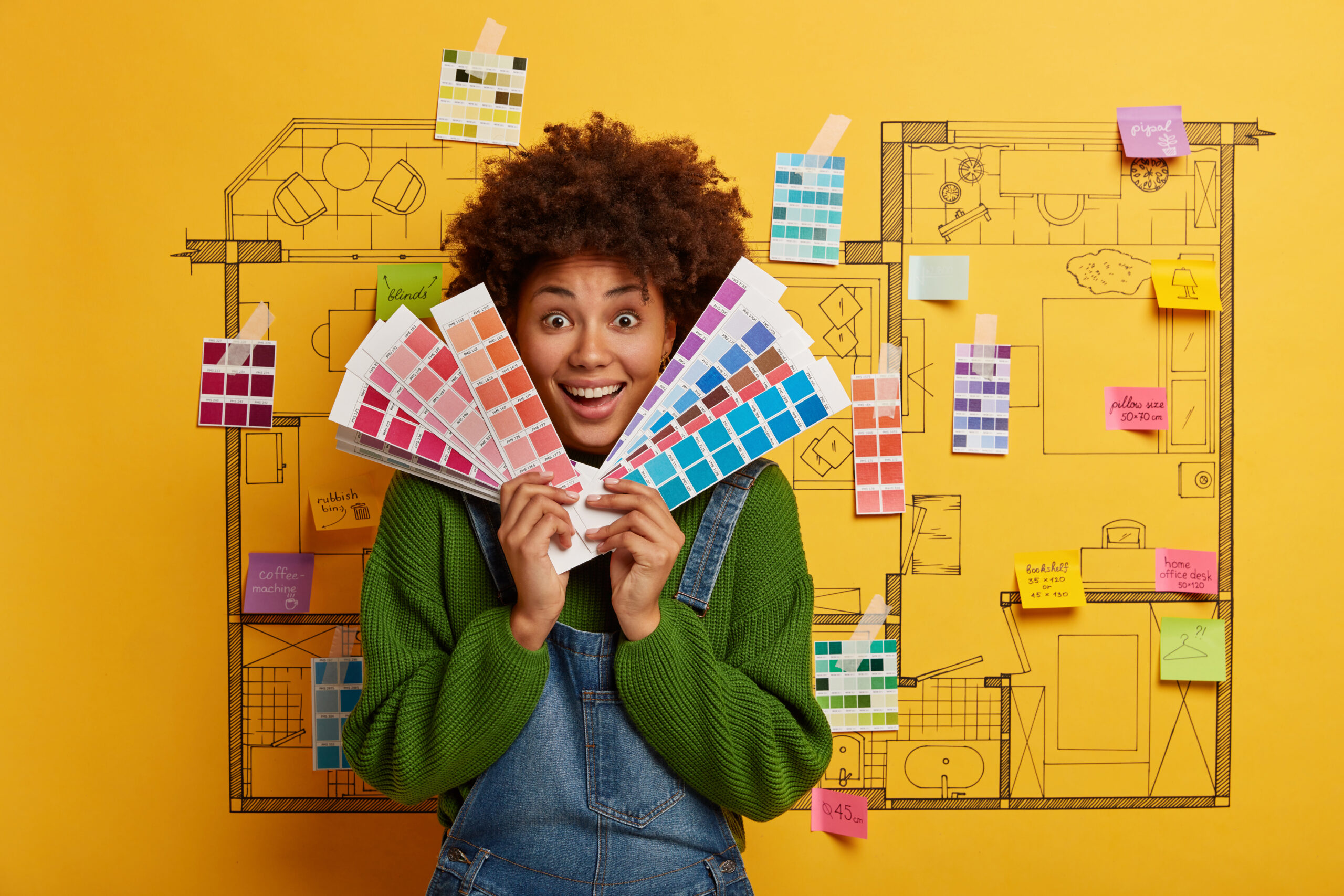 Joyful dark skinned curly woman chooses color for painting room in new house, looks through palette samples, stands against home sketch, has pleasant smile