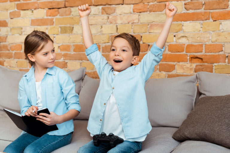 KYIV, UKRAINE - APRIL 27, 2020: sister with notebook looking at excited brother playing video game